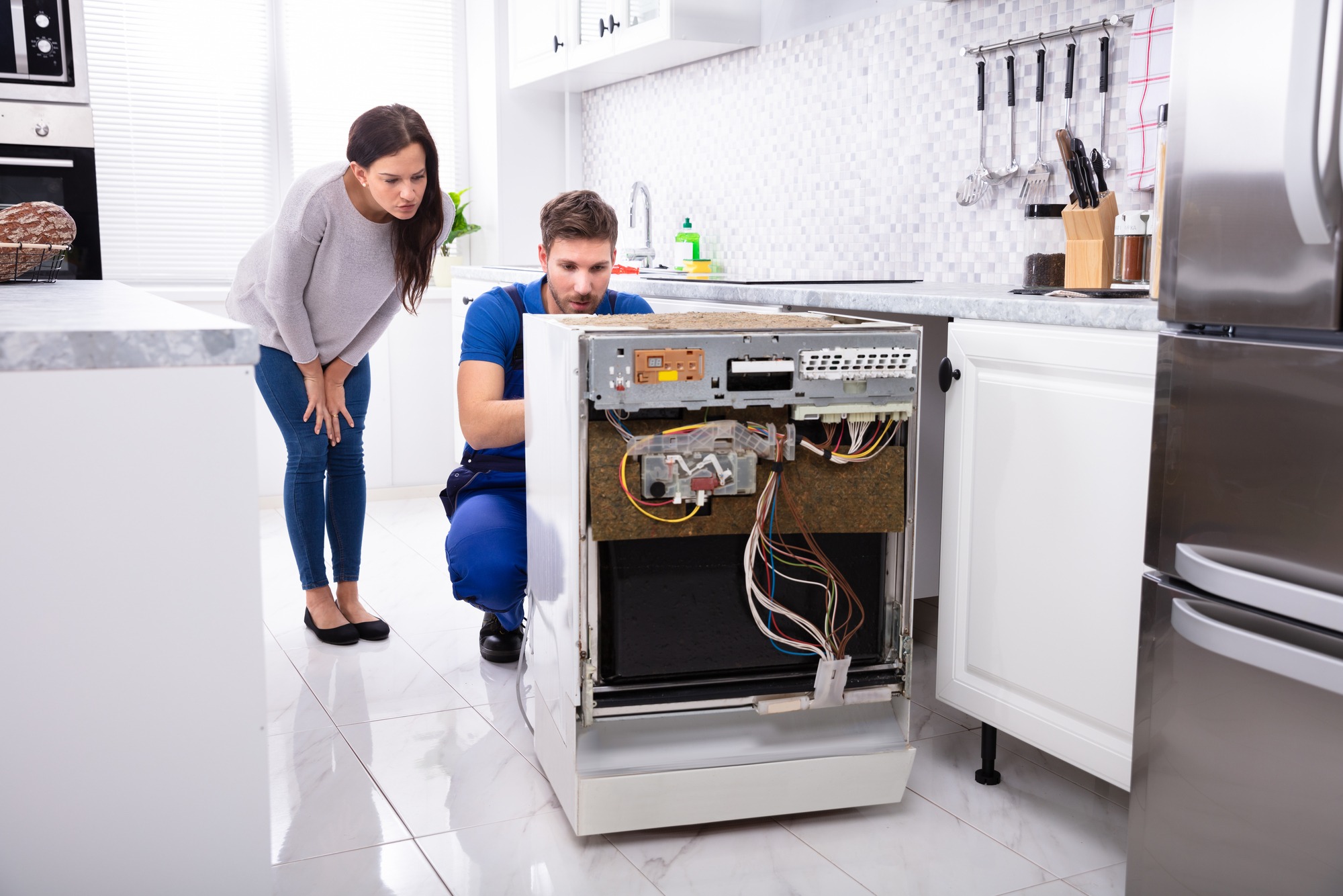 A technician inspects an open dishwasher while a concerned woman observes. The scene highlights appliance repair and maintenance in a modern kitchen.