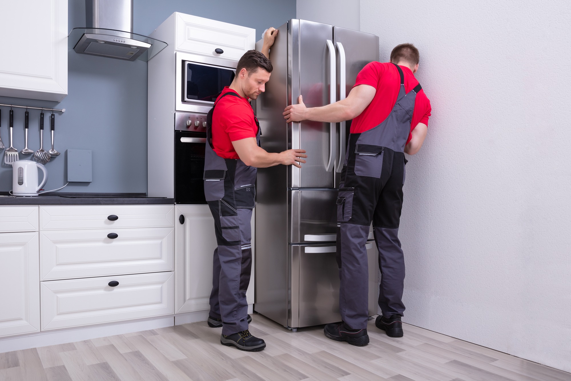 Two technicians in work attire are carefully maneuvering a stainless steel refrigerator into place in a modern kitchen.