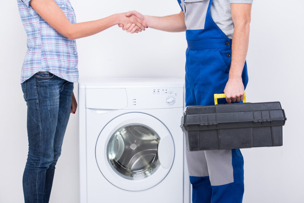 A technician in blue overalls shakes hands with a customer next to a washing machine, indicating successful service agreement.